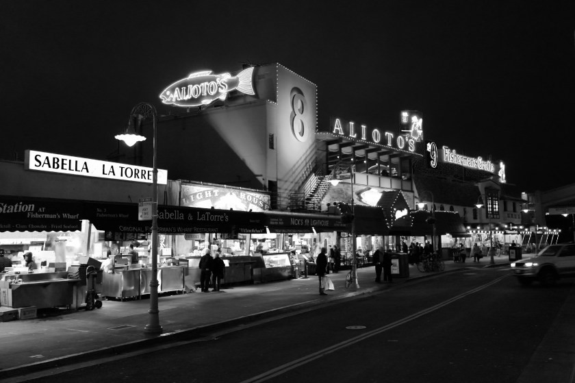 Black and white night photograph of Alioto's #8 restaurant with illuminated neon signs and marquee lights on Fisherman's Wharf in San Francisco. Food vendor stalls including Sabella LaTorre line the boardwalk in the foreground, with pedestrians visible under vintage street lamps.