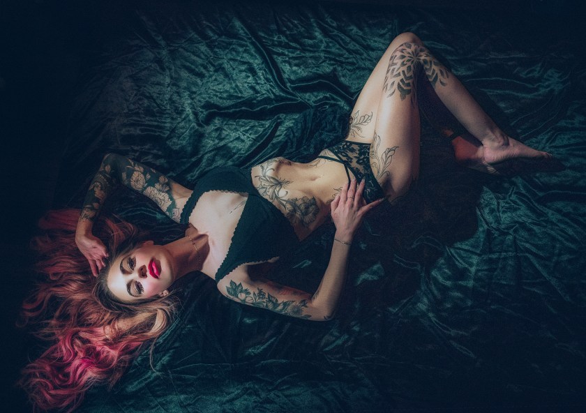 Overhead studio portrait of a tattooed woman reclining on dark fabric, wearing black lingerie under dramatic low-key lighting.