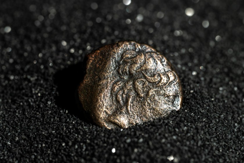 Close-up of a worn ancient Greek coin showing a raised portrait, resting on coarse black granular material.