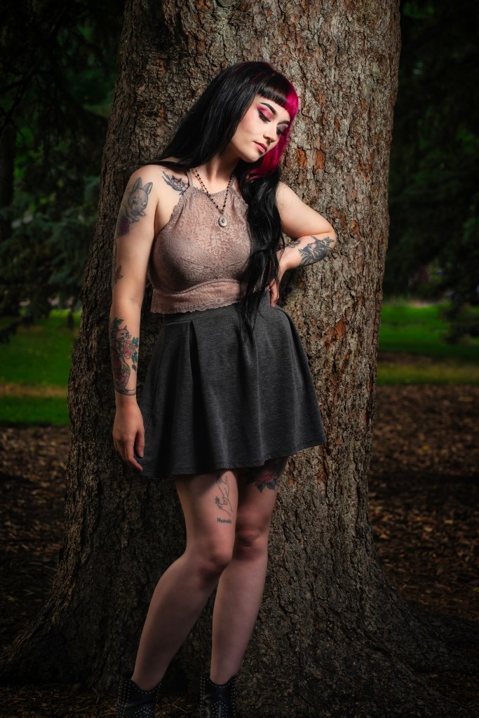 Woman with long dark hair and visible tattoos leaning against a large tree in a park, eyes closed, wearing a sleeveless top and skirt.