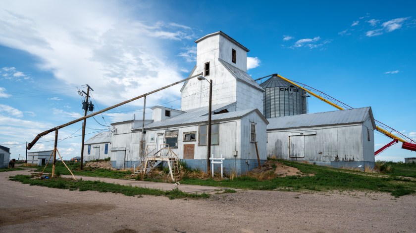 Rural grain elevator complex with white scale house buildings and metal silos under a blue sky in eastern Colorado.