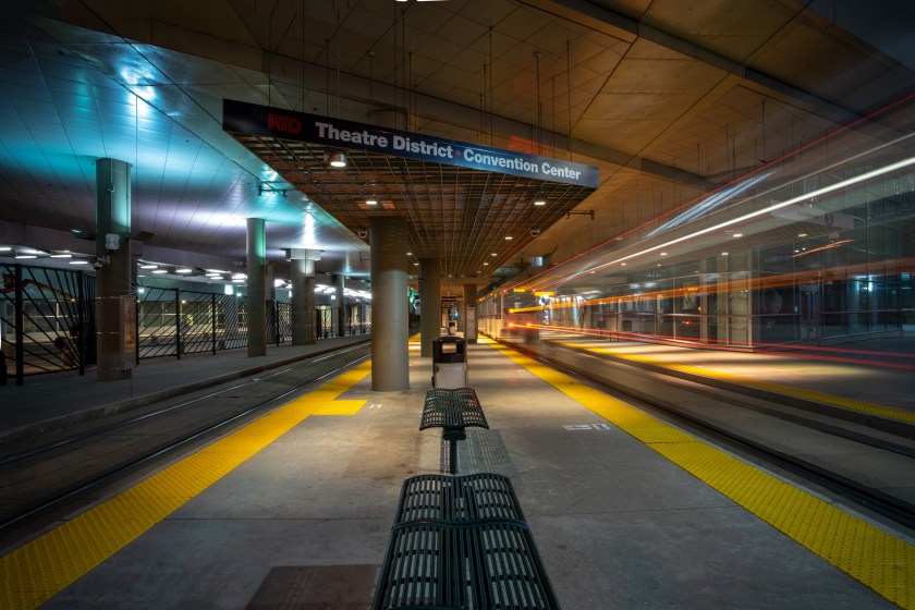 Text: Long exposure view of the RTD light rail platform at the Convention Center with blurred train lights in downtown Denver.
