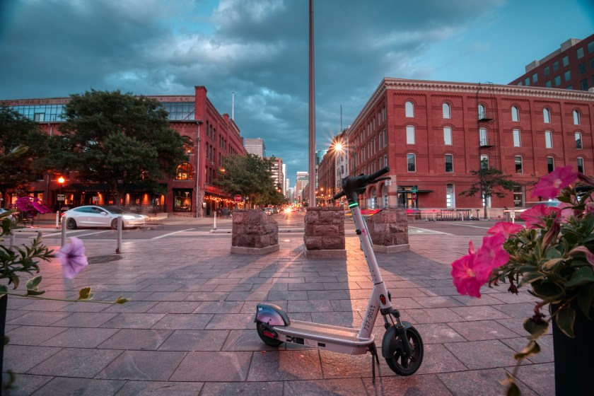 An electric scooter stands on a plaza at Denver Union Station, framed by brick buildings and evening traffic under a cloudy sky.