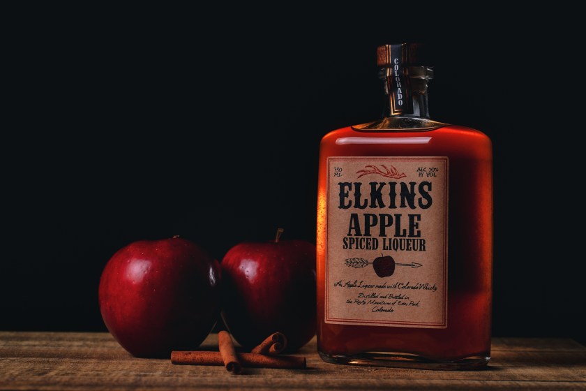 Bottle of Elkins Apple Spiced Liqueur on a wooden surface with red apples and cinnamon sticks against a dark background.