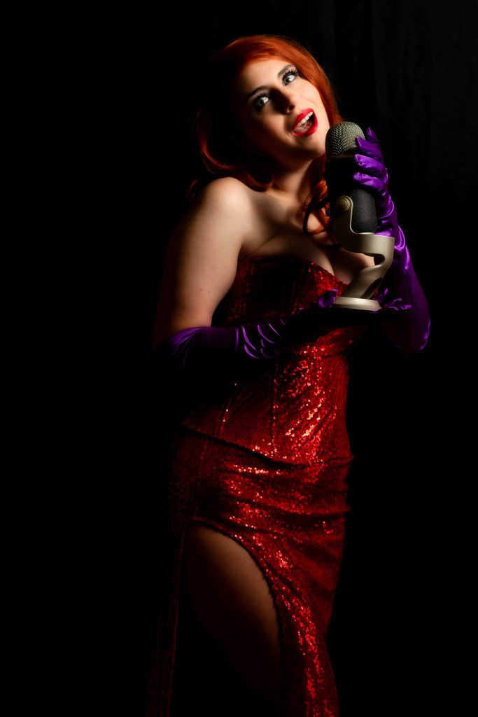 Woman in a red sequined dress and purple gloves holding a vintage-style microphone against a black background.