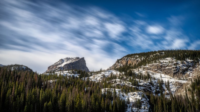 Hallett Peak rises above evergreen forest and snow-dusted slopes beneath streaked clouds in Rocky Mountain National Park.