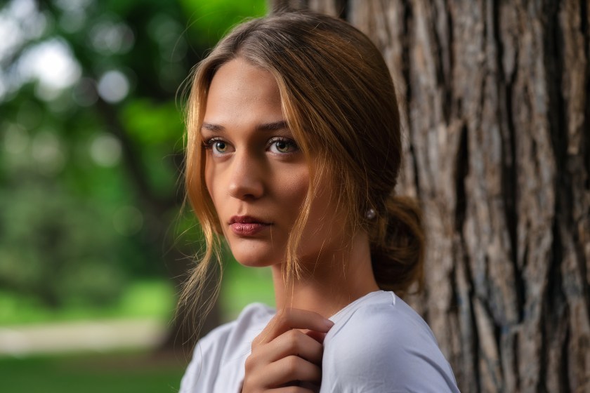 Woman with light brown hair stands beside a tree in a park, looking toward the camera with a soft green background.