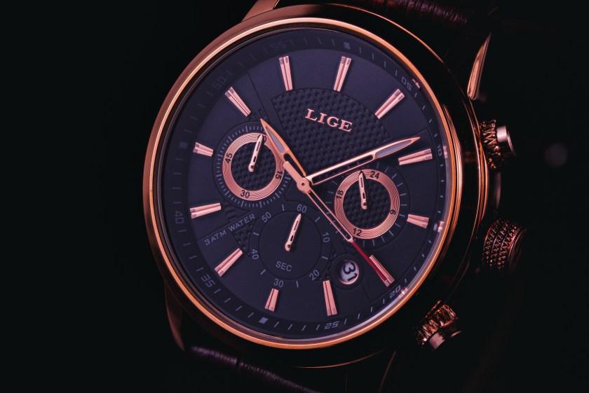 Close-up photograph of a Lige men’s chronograph wristwatch with rose gold accents on a black dial against a dark background.