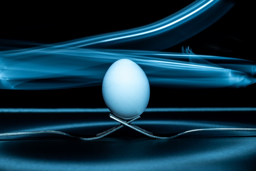 White egg balanced upright on crossed metal forks, photographed against a dark background with blue light trails created through light painting.