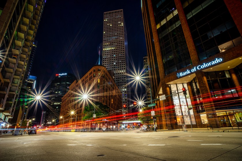 Long exposure light trails streak through a downtown Denver intersection at night, with tall buildings and streetlights surrounding the scene.