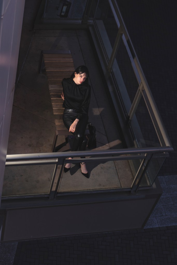 Woman dressed in black sits on a bench on an elevated walkway, viewed from above in an urban setting.