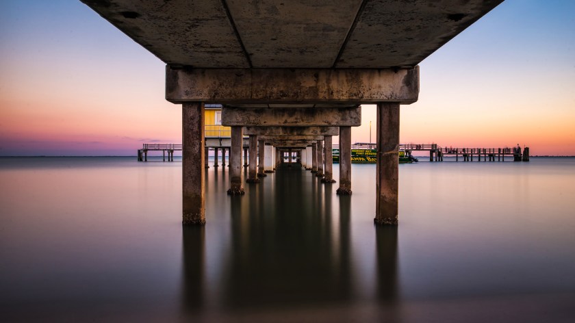 Long exposure sunset photograph taken beneath the Bay Pier at Fort De Soto Park, showing symmetrical concrete pilings and smooth reflective water.