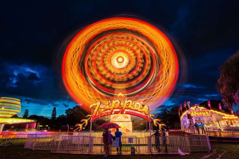 Long exposure photograph of the Zipper carnival ride at night, creating circular light trails in vivid colors with people standing below.