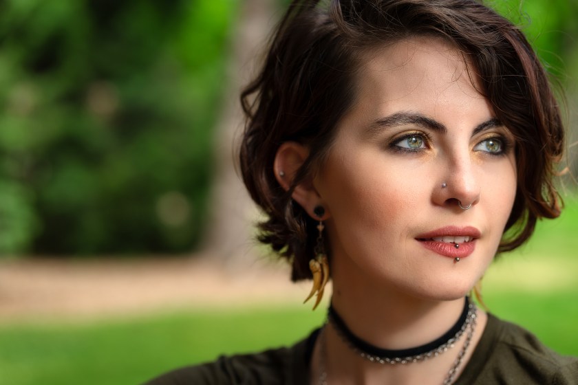 Woman with short dark hair and facial piercings looks off-camera in a park, with green foliage softly blurred behind her.