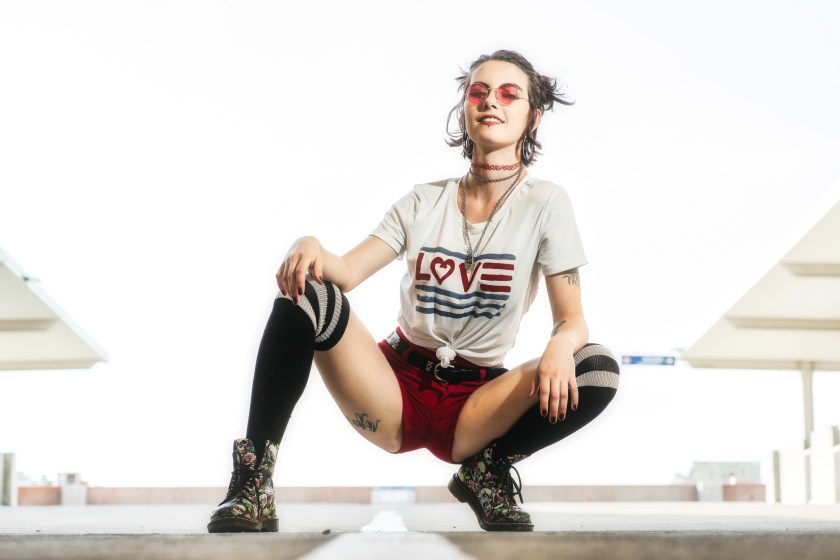 Woman wearing red shorts and knee-high socks crouches on a concrete surface in an urban setting with bright sky behind her.