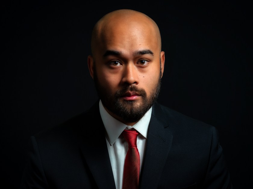 Bald man with a beard wearing a suit and red tie looks directly at the camera against a black background.