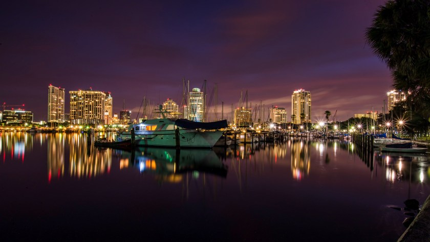 Nighttime long exposure photograph of the St. Petersburg, Florida skyline viewed from the Municipal Marina, with boats and colorful reflections on calm water.