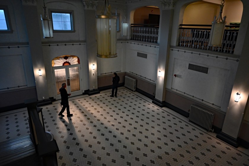 View of the Northern Hotel lobby from an upper staircase, showing patterned tile flooring, hanging lights, and two people walking across the open space below.