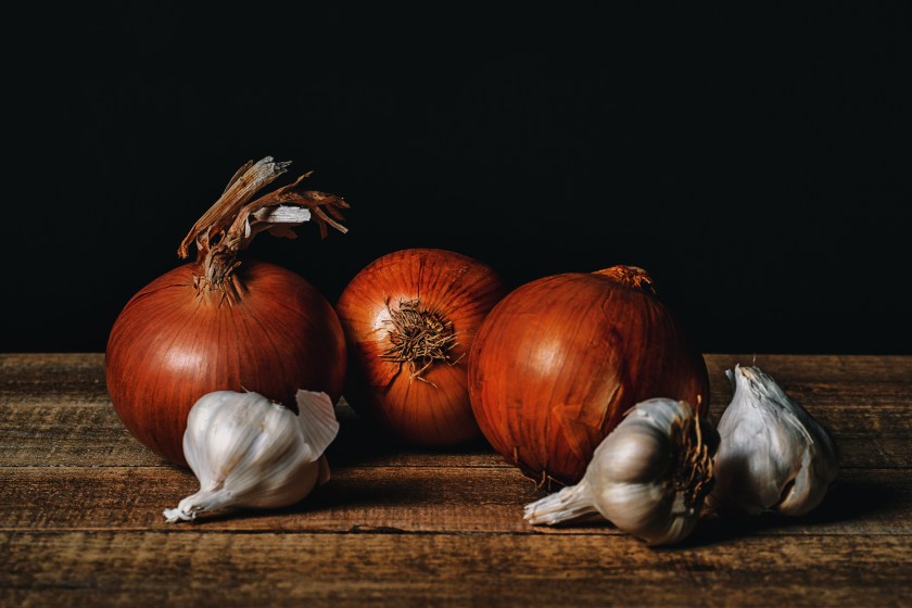 Tabletop still life of whole onions and garlic bulbs arranged on a wooden surface against a dark background.