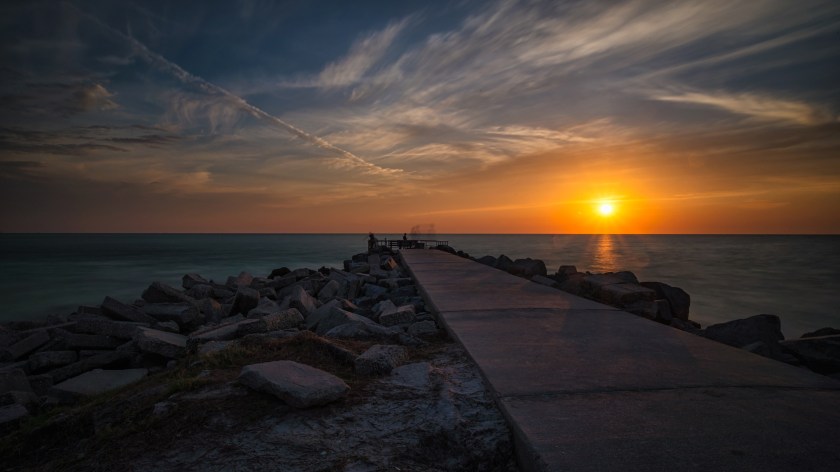 Sunset photograph of the Pass‑a‑Grille jetty in Florida, with a concrete pier extending into the Gulf of Mexico and silhouetted figures at the horizon.