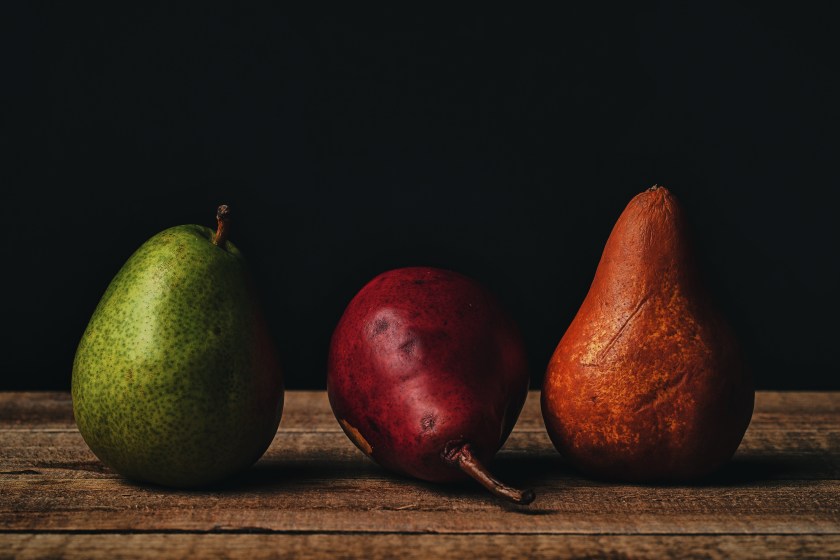 Tabletop still life of three pears arranged side by side on a wooden surface against a dark background.
