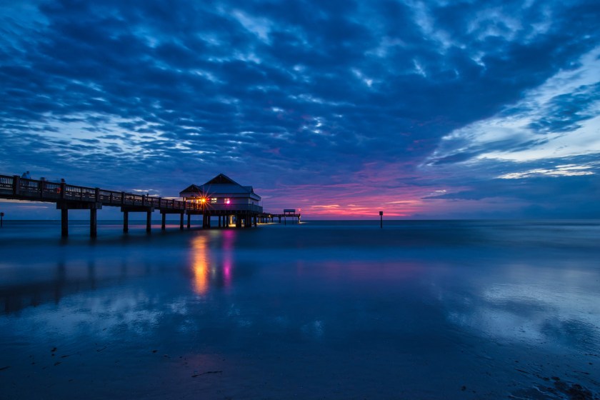 Long exposure photograph of Pier 60 on Clearwater Beach at sunset, with pink and purple sky tones reflecting on calm water.