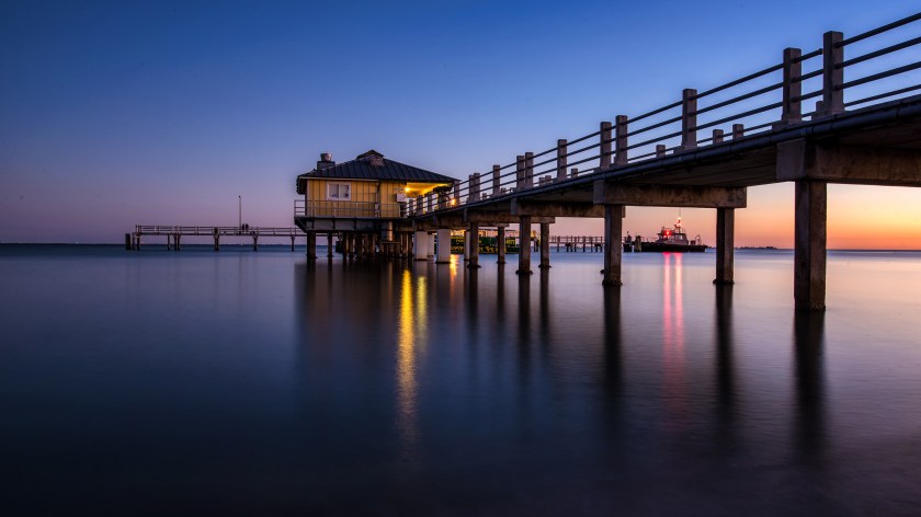 Long exposure twilight photograph of the Bay Pier at Fort De Soto Park in Florida, with a yellow pilot‑boat building illuminated over calm water.