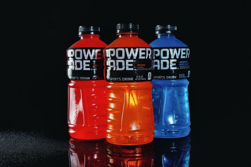 Studio still life of three Powerade sports drink bottles in red, orange, and blue on a reflective black surface.