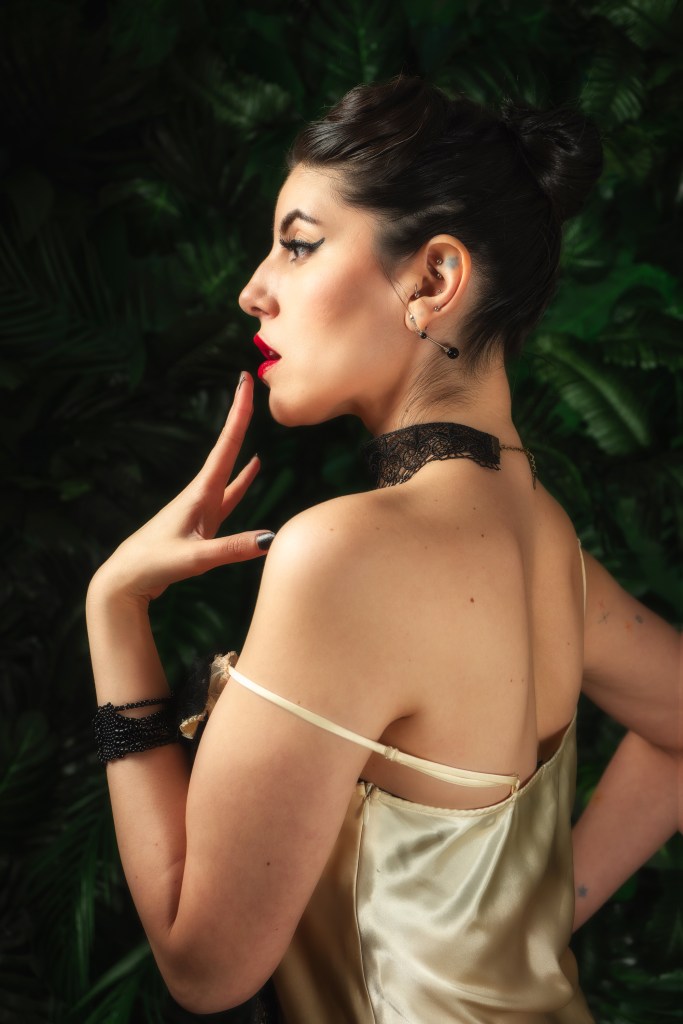 Woman in a satin dress shown in side profile against a dark leafy background, with hair in an updo and red lipstick.