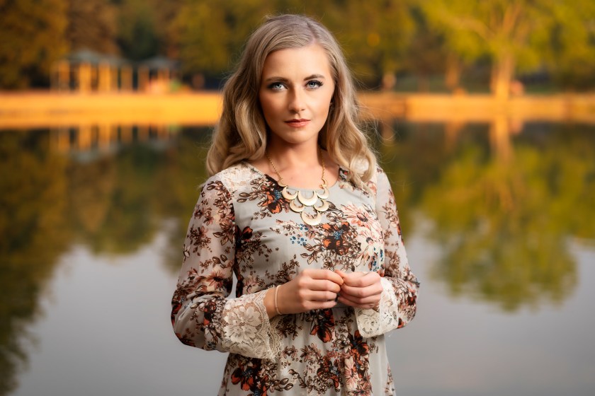 Woman in a floral dress stands by a calm lake with autumn trees reflected in the water behind her.