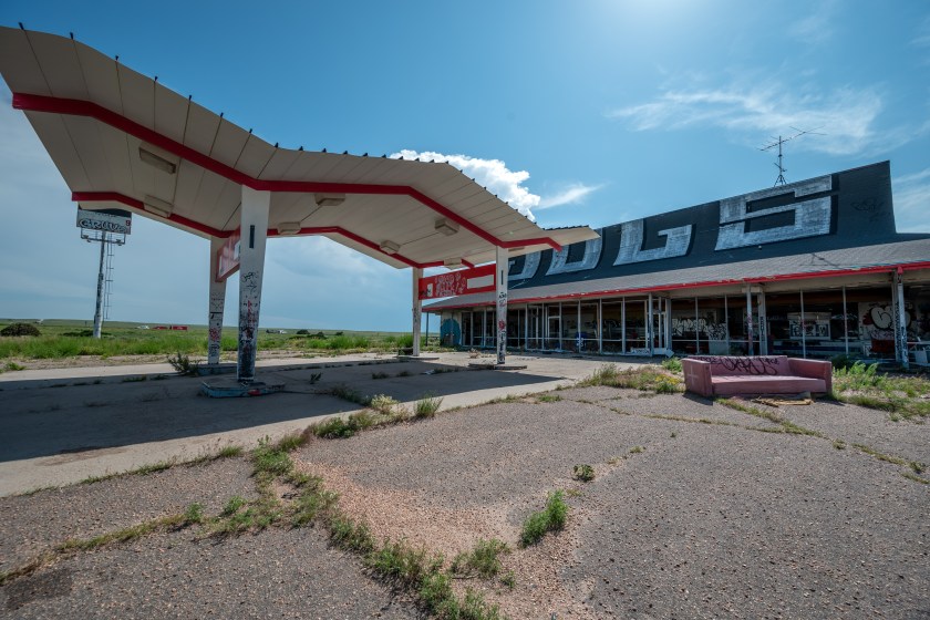 Abandoned roadside building with a former gas canopy and boarded storefront, covered in graffiti and surrounded by cracked pavement and weeds under a blue sky.
