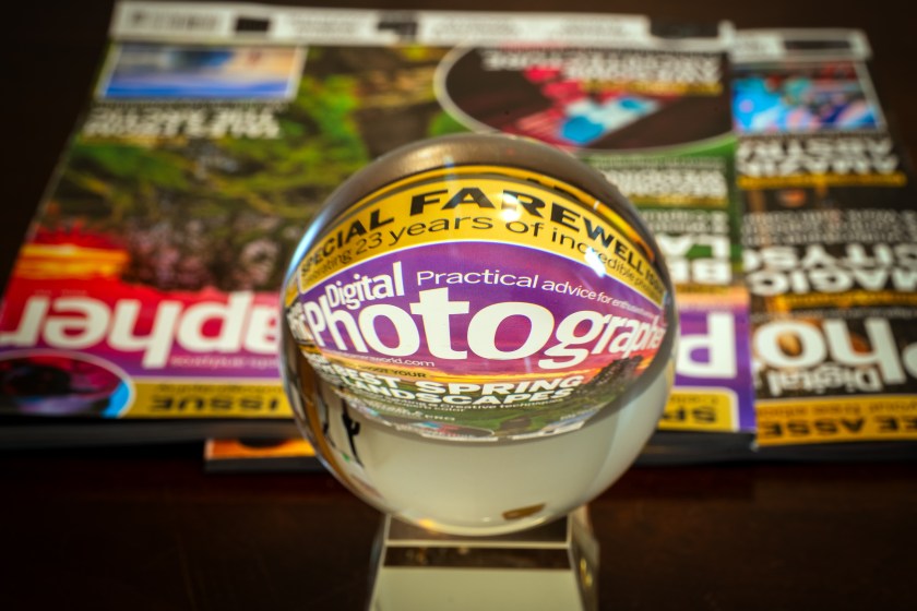 Glass sphere placed over a stack of photography magazines, magnifying and inverting the “Digital Photographer” cover through refraction.