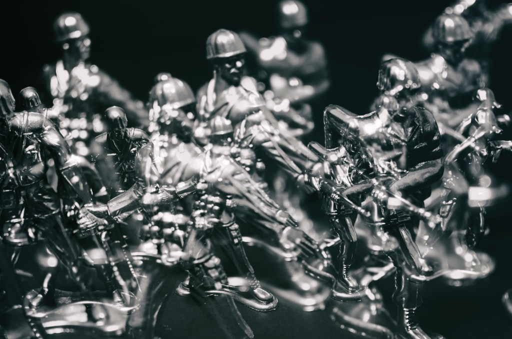 Metallic silver toy army men photographed in dramatic low light against a black background, creating an ethereal tabletop photography scene.