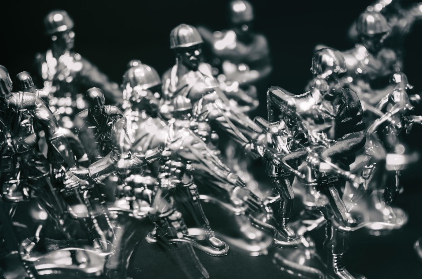 Metallic silver toy army men photographed in dramatic low light against a black background, creating an ethereal tabletop photography scene.