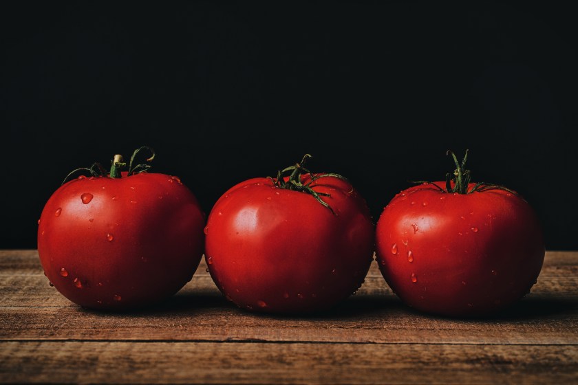Tabletop still life of three ripe tomatoes with water droplets on a wooden surface against a dark background.