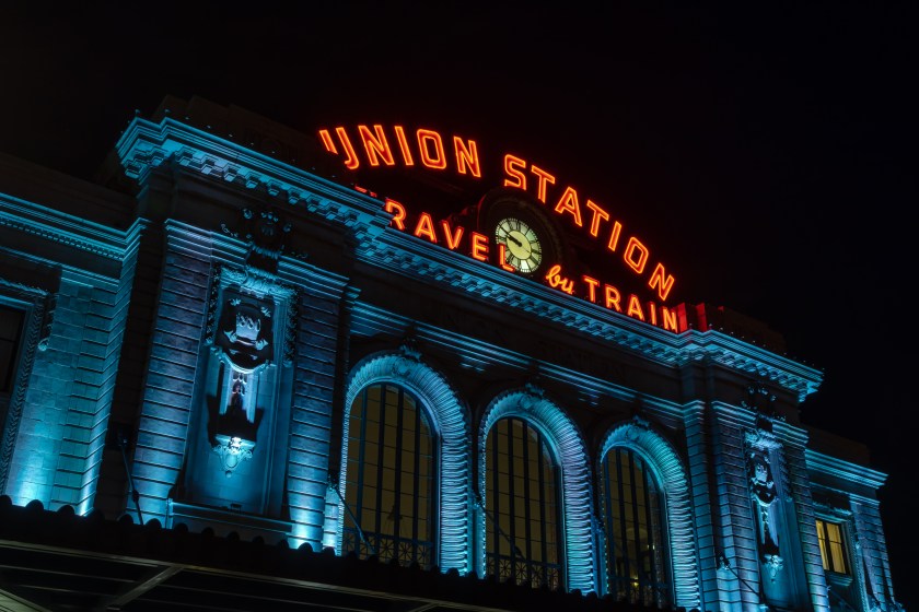 The illuminated front façade of Denver Union Station at night, with glowing neon signage and a clock above arched windows.