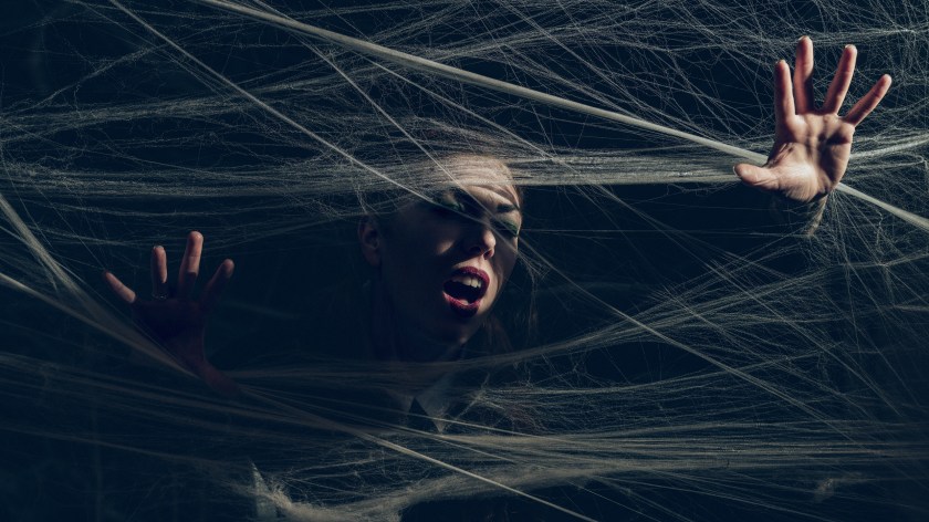 Portrait of a person pressing their hands forward through layers of stretched web-like material, with dramatic low-key lighting against a dark background.