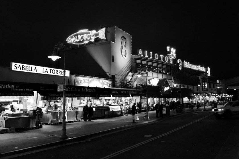 Black‑and‑white nighttime scene at Fisherman’s Wharf in San Francisco, featuring illuminated neon signs for Sabella & La Torre, Nick’s Lighthouse, Alioto’s, and Fishermen’s Grotto with people walking along the busy street.