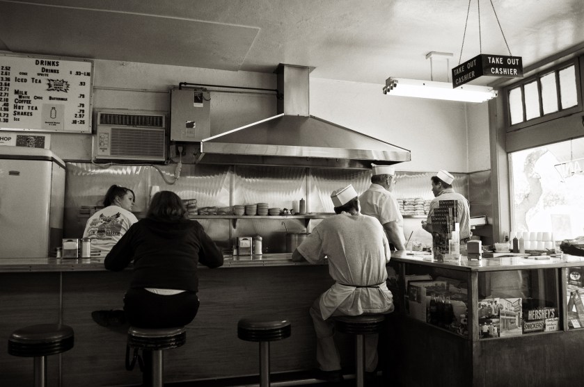Interior of the Coney Island Grill in downtown St. Petersburg, showing customers seated at the counter while three staff members in classic white diner uniforms work behind the grill beneath a vintage letterboard menu.