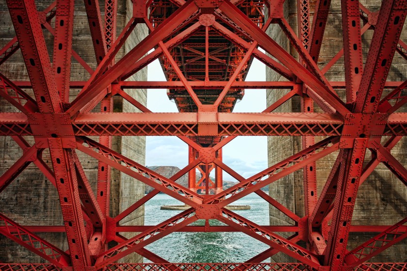 View from beneath the Golden Gate Bridge at Fort Point, showing the red steel beams, riveted joints, and geometric latticework of the bridge’s underside with concrete supports and distant hills visible beyond.